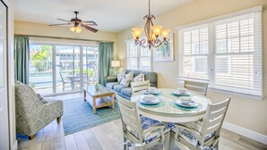 A dining area with a round glass table, white chairs, and a chandelier. There is a sofa and a window with blinds in the background. - Waterfront Retreat  (Duck Key)