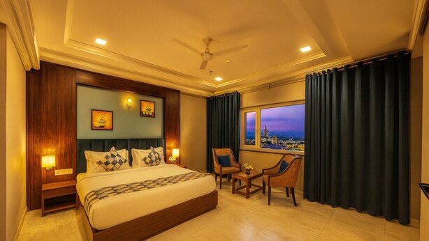 Habitación de hotel con una cama grande, dos sillones, una mesita y vistas a un paisaje urbano al atardecer.