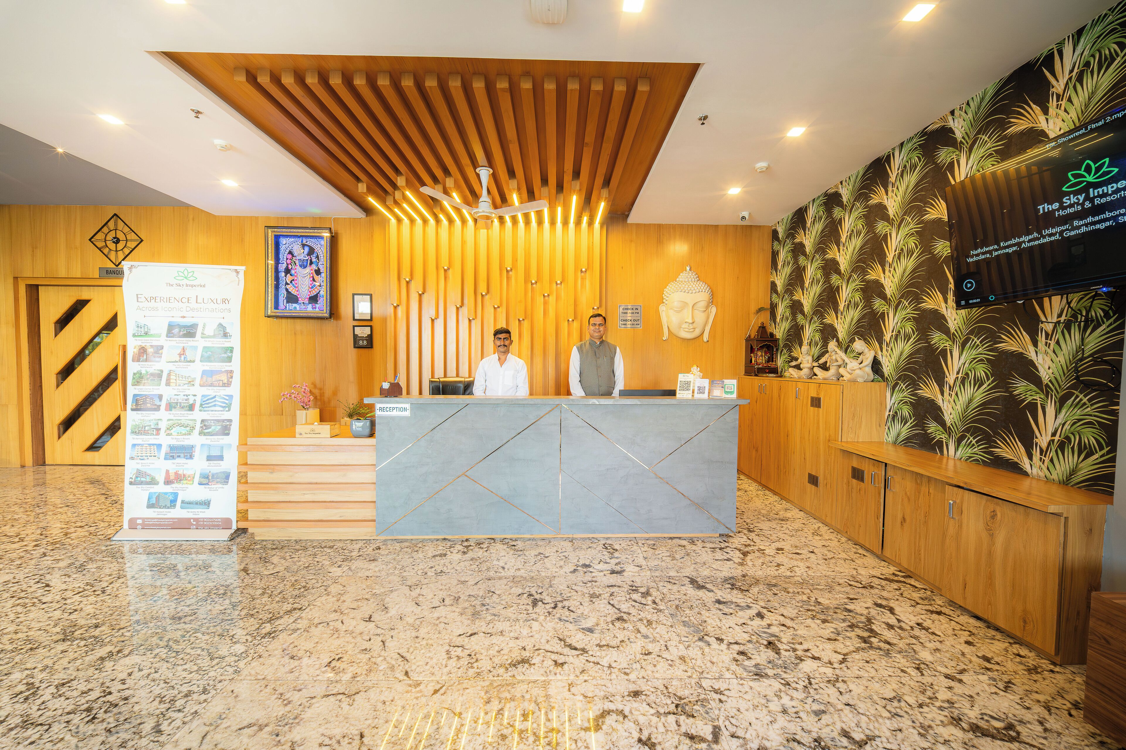 Zona de recepción de un hotel con un mostrador de madera, una estatua de Buda y una pantalla digital mostrando información del hotel.