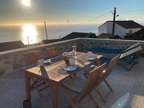 A wooden dining table set for two with a view of the sea at sunset.