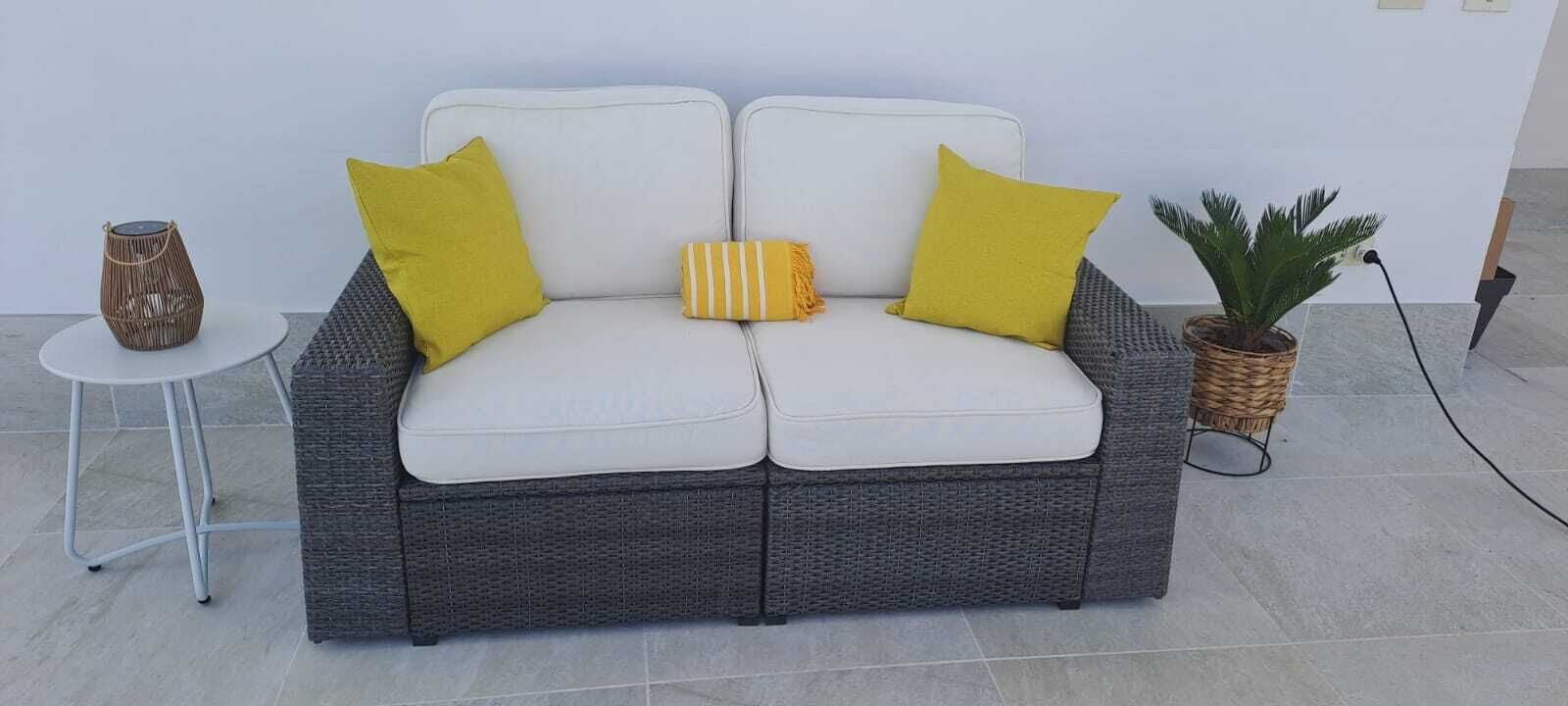 Ein graues Korbssesselsofa mit weißen Kissen und gelben Kissen, ein kleiner runder Tisch mit einem Korbkorb und eine Topfpflanze.