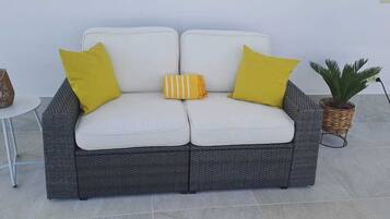 Ein graues Korbssesselsofa mit weißen Kissen und gelben Kissen, ein kleiner runder Tisch mit einem Korbkorb und eine Topfpflanze.
