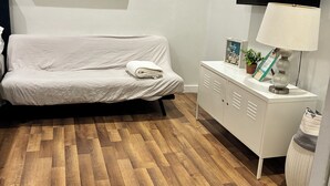 Quarto com piso de madeira, sofá branco, televisão, mesa de cabeceira com abajur e um quadro na parede.