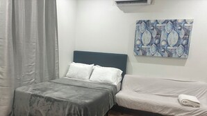 Um quarto com cama, mesa de cabeceira, manta cinza, travesseiro branco, sofá cinza e um quadro abstrato azul na parede.