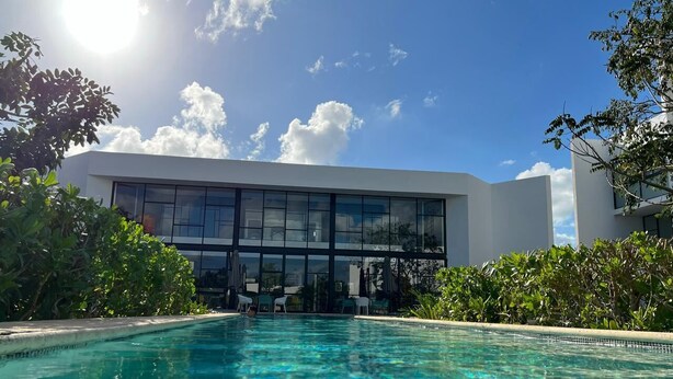 A swimming pool with clear blue water, surrounded by lush greenery and a modern building with large windows in the background. - Cozy loft with private jacuzzi. (Mérida)