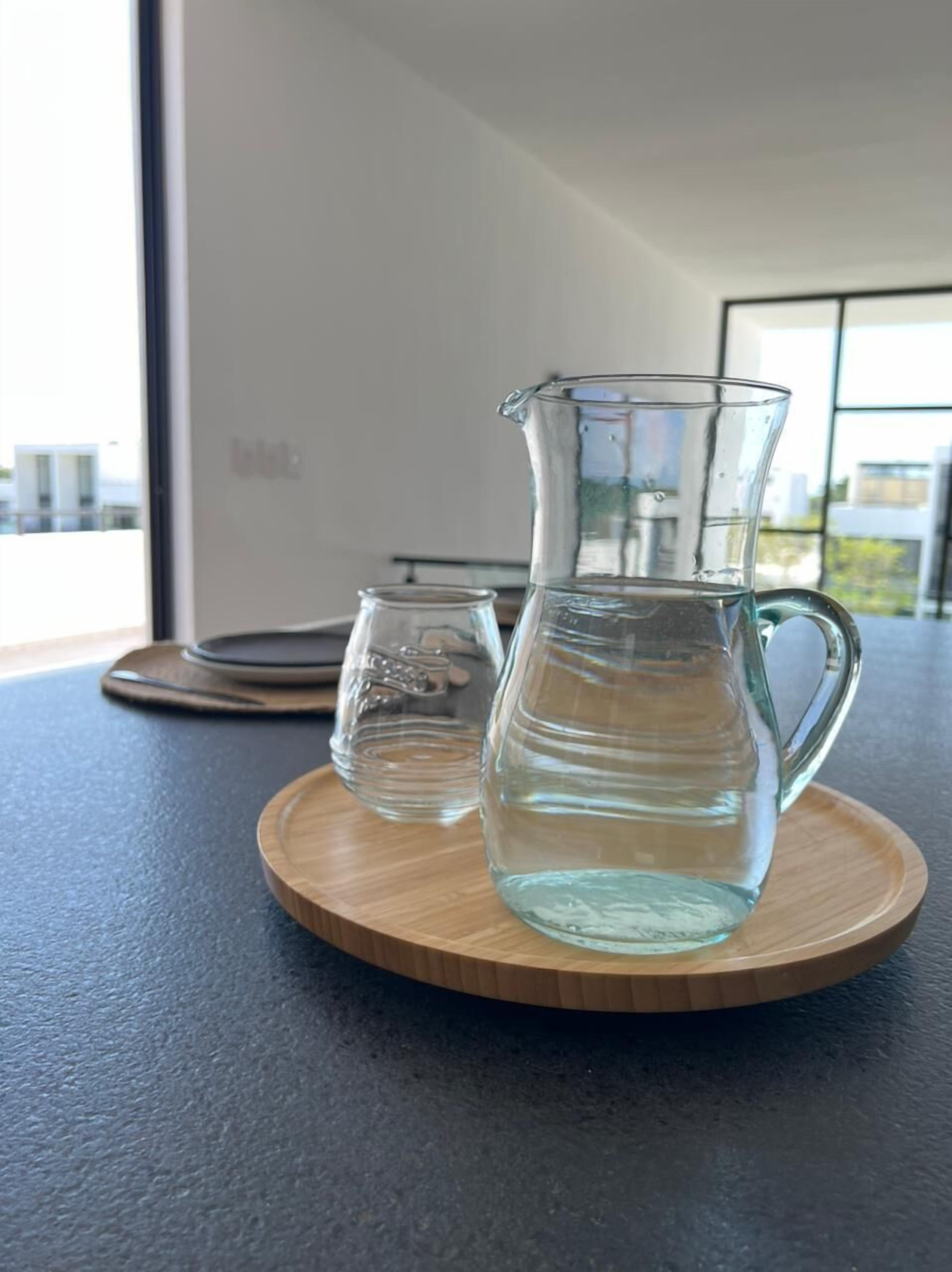 A clear glass pitcher and a smaller glass on a wooden coaster on a dark countertop.