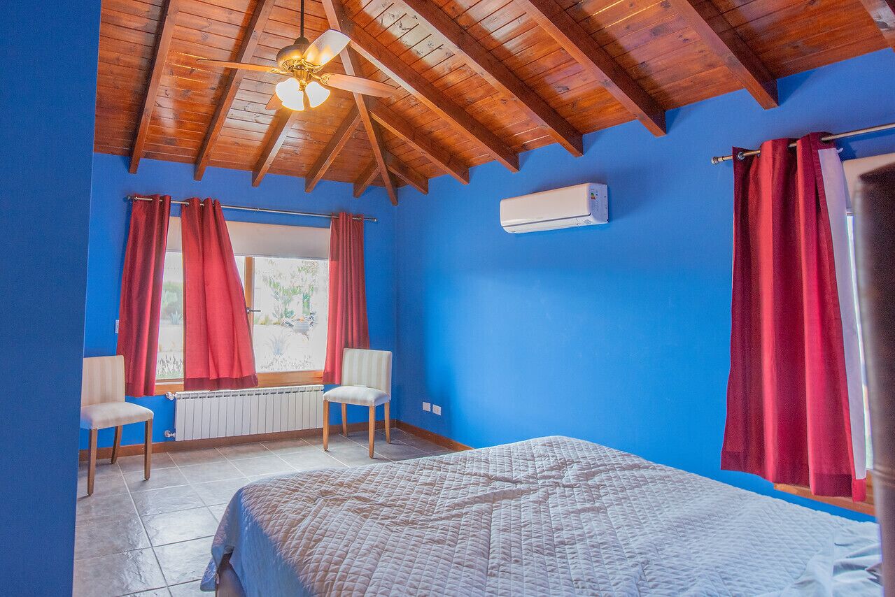 A bedroom with a wooden ceiling, a bed, two chairs, a window with red curtains, and an air conditioning unit.