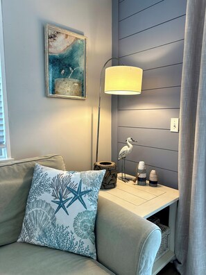 Ein Zimmer mit grauem Sofa, Kissen in Form eines Seesterns, einer Lampe, einem Wandbild und einem kleinen Tisch mit Dekorationen.