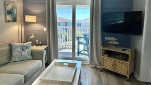 A living room with a sofa, a coffee table, a TV, and a view of the outside. - Ocean View Condo, Pool, Steps to Beach, Bikes & Beach Chairs Included (Ocean Isle Beach)