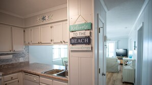 A kitchen with white cabinets, a granite countertop, and a sink. There is a sign that reads "Welcome to the Beach" with a relaxing message. - Ocean View Condo, Pool, Steps to Beach, Bikes & Beach Chairs Included (Ocean Isle Beach)