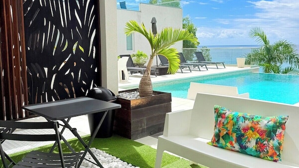 A modern outdoor lounge area with a pool, a white sofa with a colorful pillow, a round table, and a decorative plant. - Cosy (Apt7) - Patio, Sea View and pool (indigo Bay)