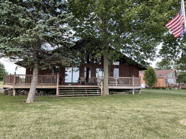 A wooden cabin with a deck, surrounded by trees and a grassy lawn.