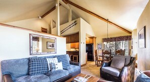 A cozy living room with a sofa, armchair, and coffee table, featuring a wooden ceiling and a dining area with chairs and a table.