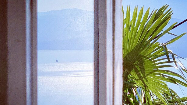 Blick durch ein Fenster auf ein Gewässer mit Bergen im Hintergrund.