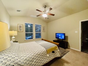 A bedroom with a bed, a ceiling fan, a TV, and two windows with blinds.