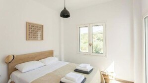 Un dormitorio con cabecera de madera, cama con ropa blanca, lámpara colgante, ventana con vista a la vegetación y una cita enmarcada en la pared.