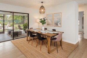 A dining area with a wooden table, black and pink chairs, a hanging light fixture, and a view through glass doors.