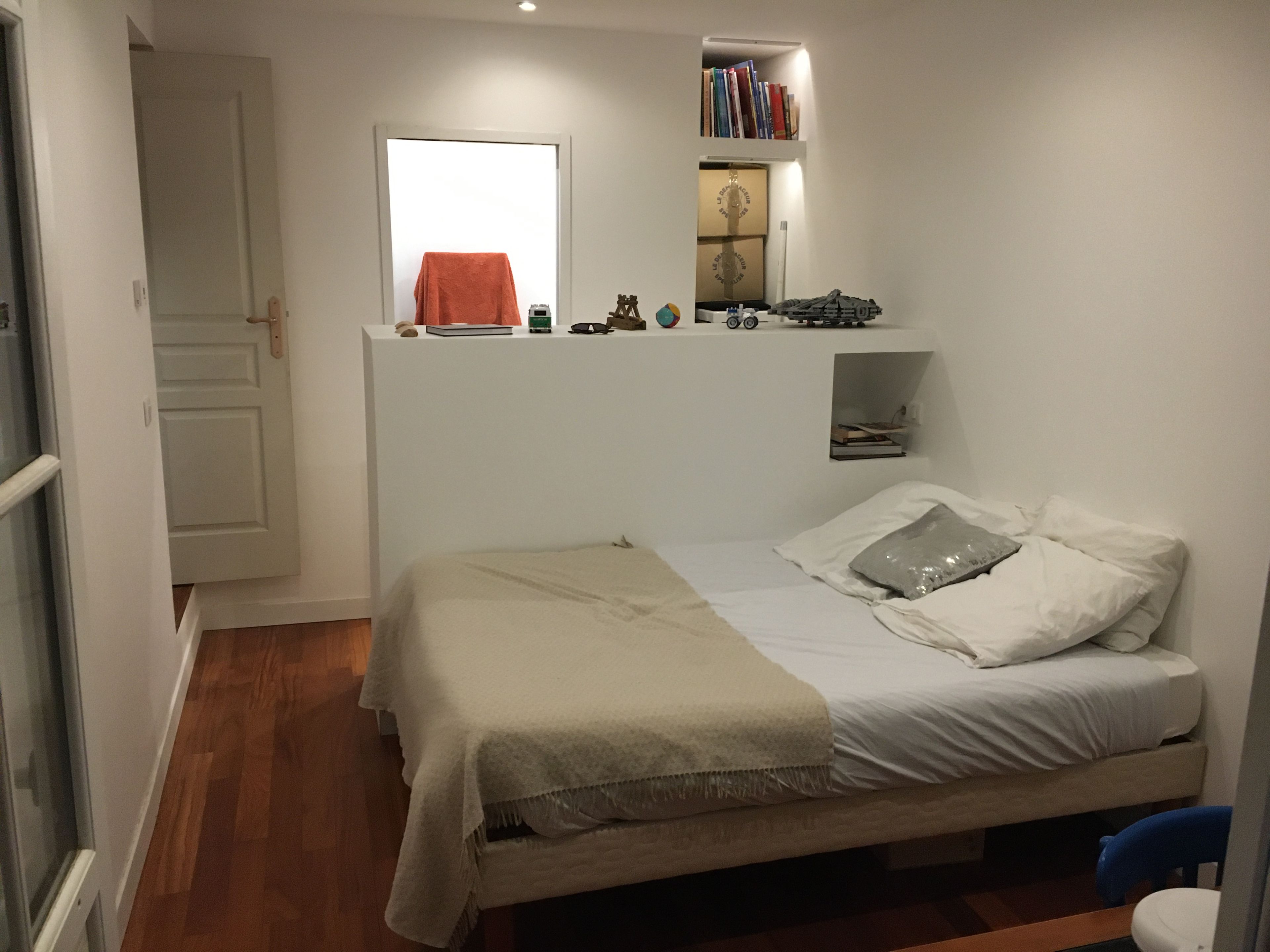 Une chambre à coucher avec un lit, une petite étagère avec des livres et un jouet, une fenêtre et une porte.