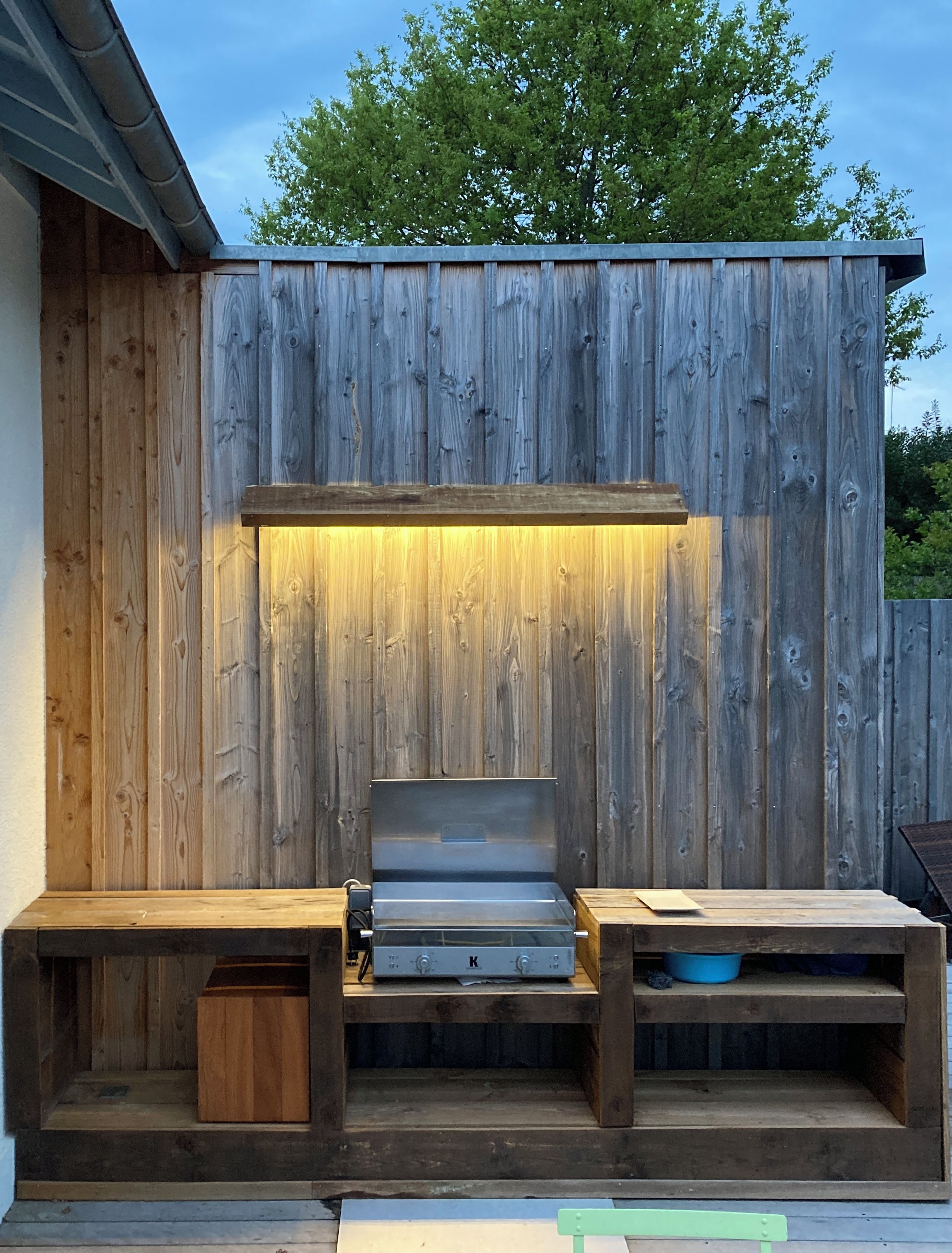 An outdoor grill area with a wooden backdrop, a metal grill, and wooden benches.
