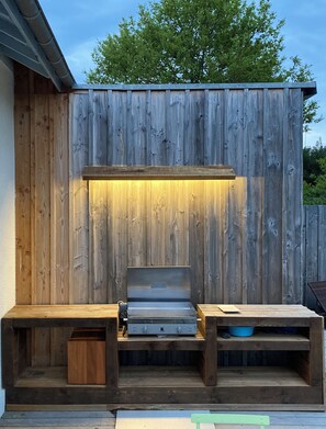 An outdoor grill area with a wooden backdrop, a metal grill, and wooden benches. - The Agape in Andernos, a 4-star hotel (Andernos-les-Bains)