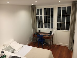 Ein Schlafzimmer mit einem Bett, einem Schreibtisch und einem Stuhl. Es gibt ein Fenster mit Vorhängen, eine Heizung und ein an der Wand befestigtes Regal.
