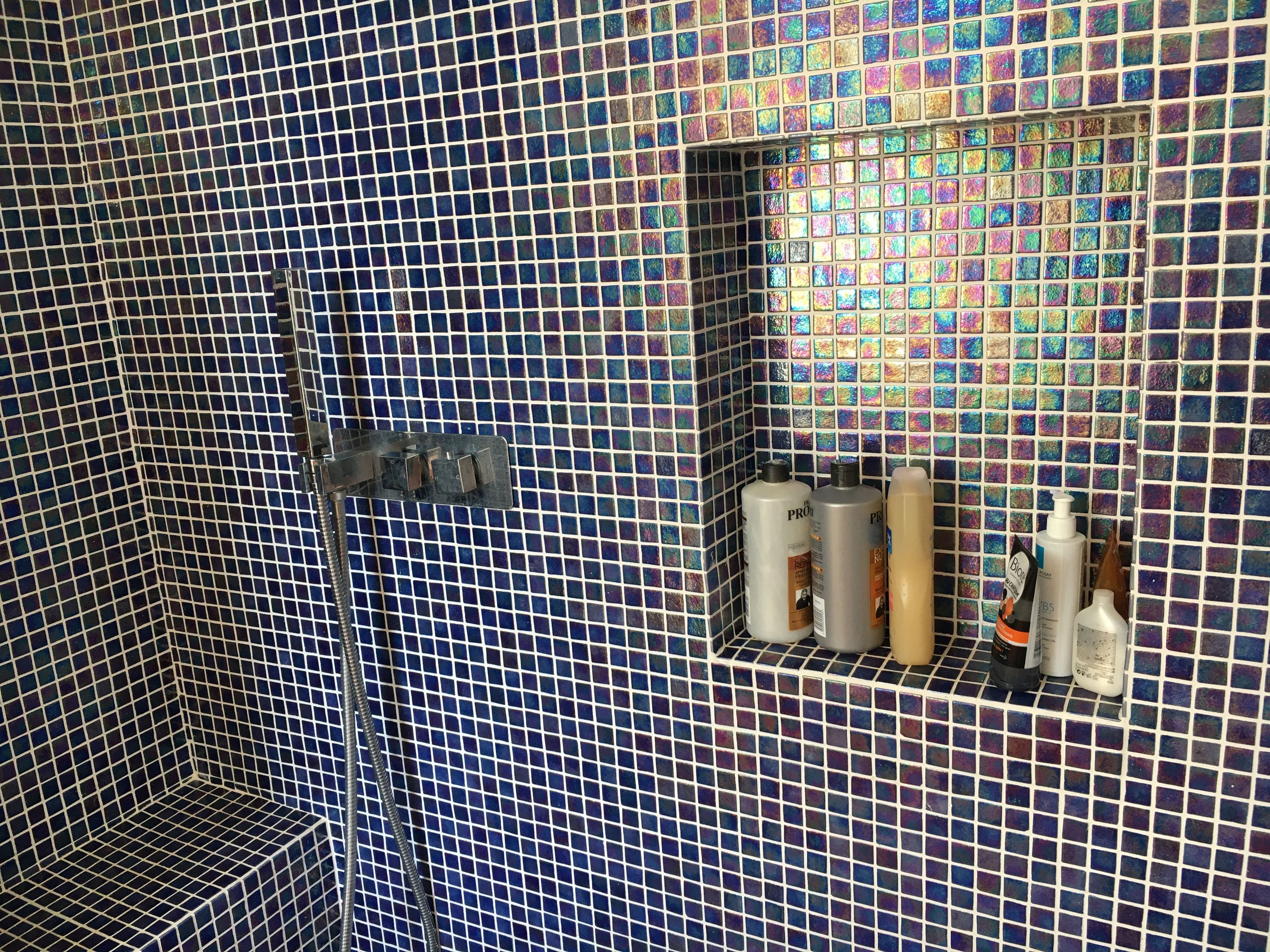 Une salle de bain avec un sol et des murs carrelés, un espace douche et une étagère contenant des articles de toilette.