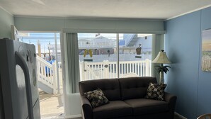 A living room with a brown sofa, a lamp, and a view of a balcony with a white fence and a building in the background. - The Townhouse Cozy Ocean-front first floor condominium. (Wildwood Crest)