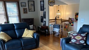 Una sala de estar con dos sofás de cuero azulados, suelo de madera, una escalera y una pared decorada con marcos y un cartel.
