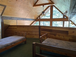 Uma cabana com duas beliches de madeira, teto inclinado com ventilador e janelas amplas com vista para árvores.