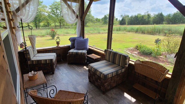 A covered outdoor seating area with wicker furniture, cushioned chairs, and a view of a grassy field. - Riverside "Barndominium" w/ easy access to water (Eureka Springs)