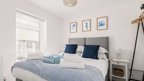 A neatly made bed with a striped blanket, a bedside table with a lamp, and framed artwork on the wall. - Seas the Day - Moments from Hamworthy Beach (Poole)