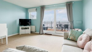 A modern living room with a flat-screen TV, a white sofa, and a balcony with a table and chairs. - Ocean Breeze - Two Bedroom Apartment with Parking (Poole)