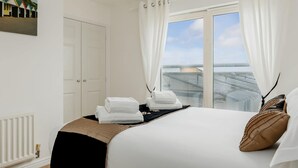 A hotel room with a bed, white curtains, a view of the sea, and a painting on the wall. - Ocean Breeze - Two Bedroom Apartment with Parking (Poole)