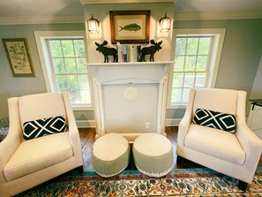 Una sala de estar acogedora con dos sillones blancos, una chimenea con un cuadro de un pez encima, y dos mesitas con cojines decorativos.