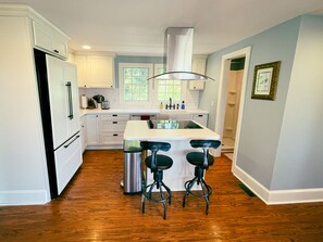A modern kitchen with white cabinets, a stainless steel refrigerator, a central island with bar stools, and a window overlooking greenery.