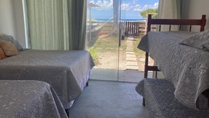 A room with two beds, a wooden headboard, and a view of a garden and ocean through a glass door. - Feet in the Sand. (Arraial do Cabo)