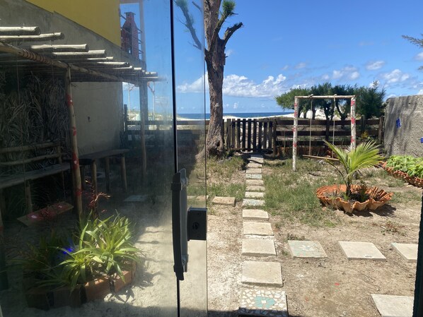 A courtyard with a stone pathway leading to a wooden gate, surrounded by potted plants and a view of the ocean. - Feet in the Sand. (Arraial do Cabo)