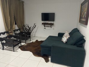 A living room with a sofa, a flat-screen TV mounted on the wall, a wooden shelf, and a brown leather rug. - Feet in the Sand. (Arraial do Cabo)