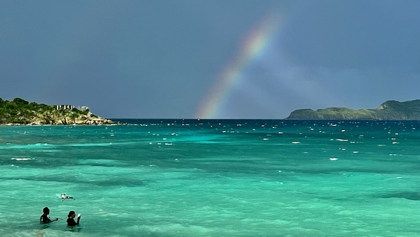 A beach with clear turquoise water, a rainbow, and a distant island with buildings.