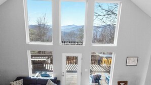 A spacious living area with a high ceiling, large windows offering mountain views, a dark-colored sofa, a wooden coffee table, and a dining table with chairs. -  Long Range Mtn View Hottub Game Room Firepit Grill Fireplace close to Asheville (Fairview)