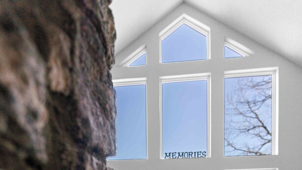 A spacious living room with a stone wall, a blue sofa, a wooden coffee table, and a black ottoman. There are large windows with a "MEMORIES" sign, and a deck is visible in the background. -  Long Range Mtn View Hottub Game Room Firepit Grill Fireplace close to Asheville (Fairview)