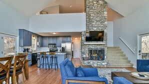A spacious living area with a stone fireplace, a kitchen with blue cabinets, and a dining area with wooden chairs. -  Long Range Mtn View Hottub Game Room Firepit Grill Fireplace close to Asheville (Fairview)