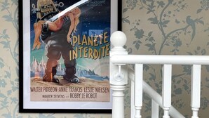Une affiche de film encadrée, représentant le film « Planète interdite », est accrochée à un mur recouvert d’un papier peint à motifs floraux.