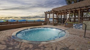 Uma banheira de hidromassagem com um deck de madeira e área de estar coberta, com vista para uma praia com guarda-sóis.