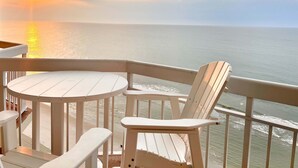 Varanda com mesa branca e duas cadeiras, com vista para o mar tranquilo ao pôr do sol.