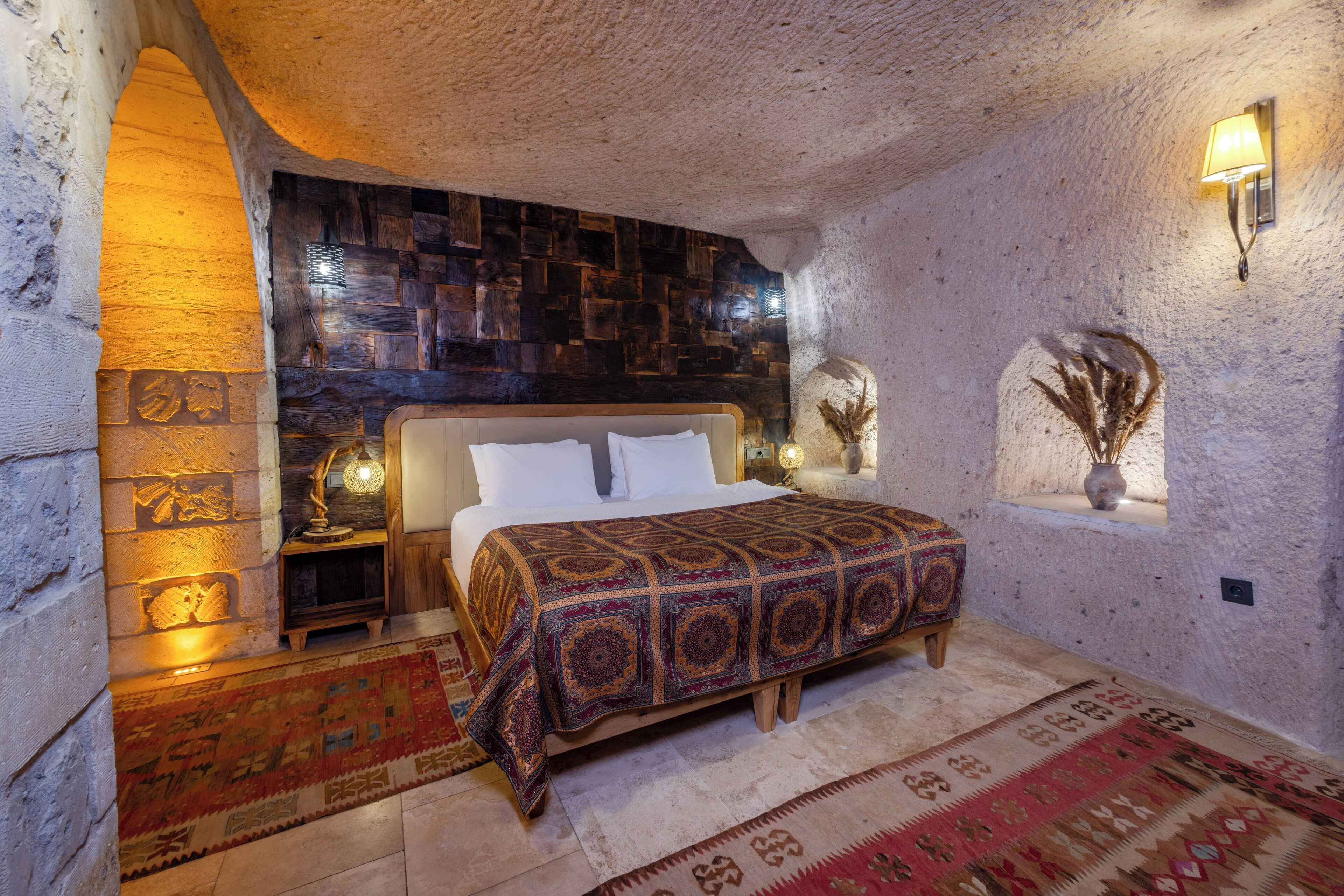 Ein Schlafzimmer mit einer Steinwand, einem Holzkopfteil, einer gemusterten Tagesdecke und einem Nachttisch mit Lampe.