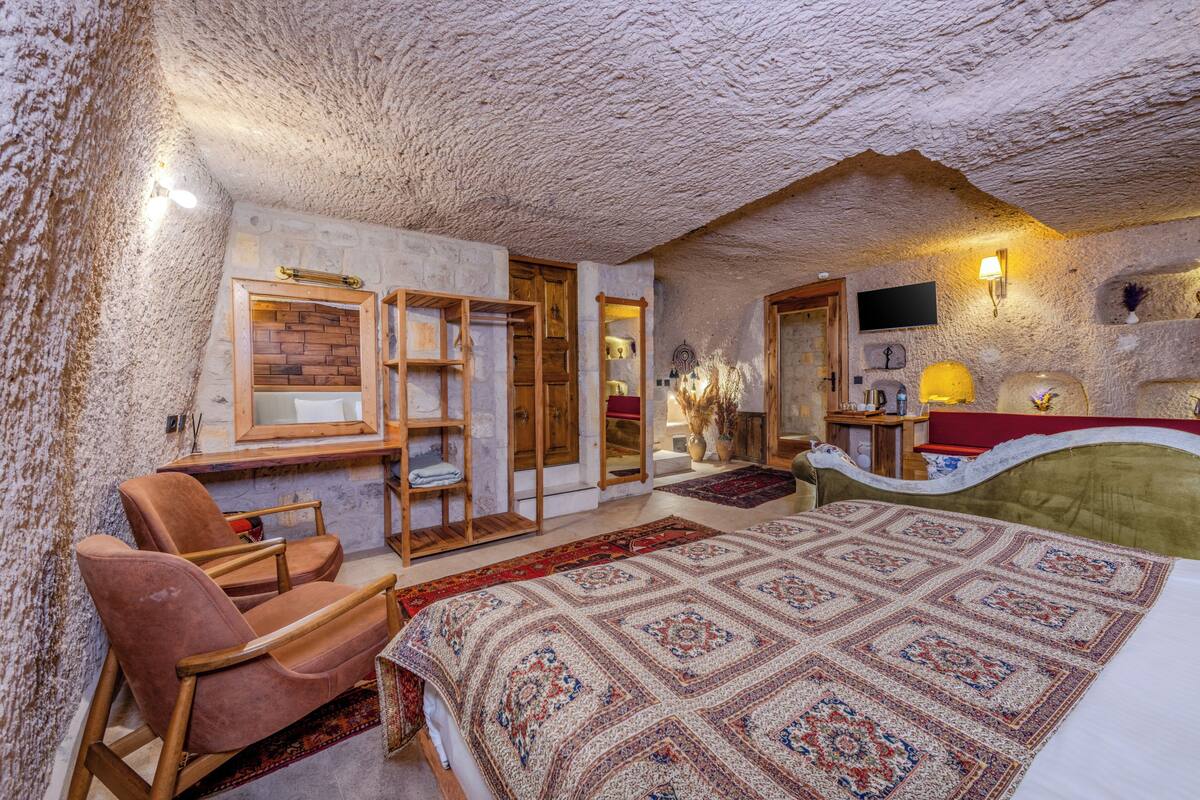 Een slaapkamer met een rustiek stenen plafond, een groot bed met een geborduurde deken, een houten stoel en een klein tafeltje met een lamp.