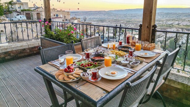 A table set for breakfast on a balcony with a scenic view, including hot air balloons in the distance.