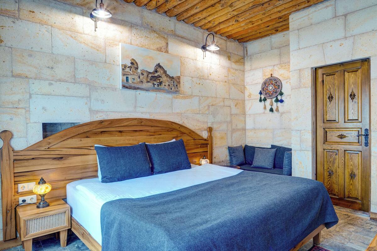 Een slaapkamer met een houten hoofdbord, een bed met blauw beddengoed, een nachtkastje met een lamp, en een stenen muur met een schilderij en een dreamcatcher.