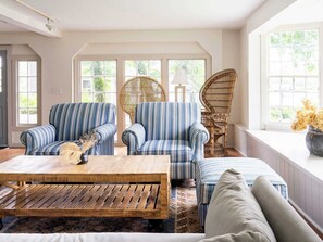 A room with a large window, two striped armchairs, a rattan chair, a wooden coffee table, and a patterned rug.